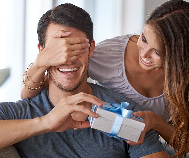 Woman covering the eyes of her spouse and handing him a present