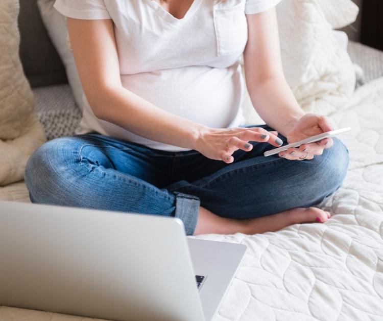 Pregnant woman scrolling on her phone with her laptop open in front of her