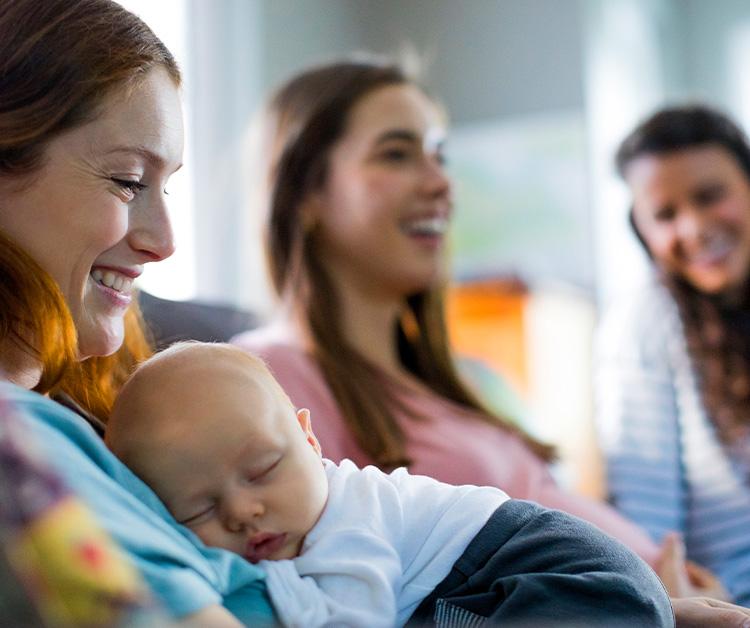 Three women hanging out and one of the women has a sleeping baby on their chest