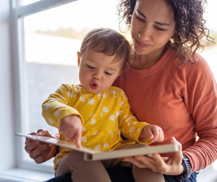 mother and child read a book together by a window. The child points at the book, wearing a yellow floral dress.